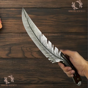 May include: A decorative knife with a feather-shaped blade. The blade is silver with etched lines, and the handle is dark wood. The knife is held against a dark wood background. The text "ARKOBLADESCO" is visible.