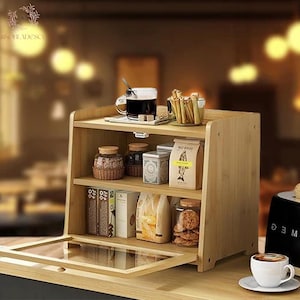 May include: A light brown bamboo bread box with three shelves. The top shelf holds a cup of coffee, a small dish of breadsticks, and a sugar cube. The middle shelf has jars and a tin. The bottom shelf has jars, bags, and a glass door.