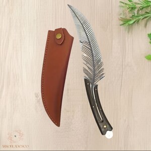 May include: A decorative knife with a feather-shaped blade and a dark wooden handle. The blade is silver and detailed. A brown leather sheath with a snap closure is included. The knife is designed for display or collection.