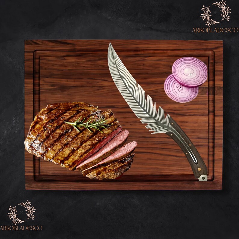 May include: A dark wooden chopping board features a sliced steak, red onion rings, and a decorative knife. The knife has a feather-shaped blade and a dark wooden handle. A sprig of rosemary sits atop the steak.