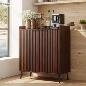 May include: A dark brown wooden cabinet with vertical ribbed detailing on the doors. The cabinet has black legs and is set against a light wood-paneled wall. A coffee machine, bottles, and decorative items sit on top.