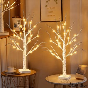 May include: Two white birch tree decorations with warm white LED lights. The trees have a textured bark design and are mounted on square white bases. The trees are on small round tables.
