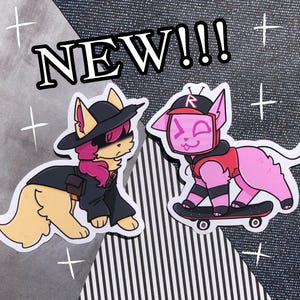 Puede incluir: Dos pegatinas de gatos de dibujos animados. Uno es beige y negro, con sombrero y abrigo. El otro es rosa, con chaleco rojo y gorra, montando en monopatín. La palabra "NEW!!!" está arriba.