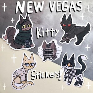 May include: A selection of "New Vegas Kitty" stickers. The stickers depict cartoon cats in different costumes, such as a hooded cloak, a gas mask, and a lab coat. The word "Stickers!" is also visible.