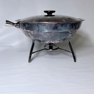 May include: A silver-toned chafing dish with a lid and a black handle. The dish sits on a three-legged stand with a circular support. The lid has a black handle. The chafing dish is designed for keeping food warm.
