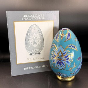 May include: A decorative egg with a turquoise background and gold and blue floral design. The egg is on a gold base. A card with the text "The Collector's Treasury of Eggs" and "Turkish Tradition" is in the background.