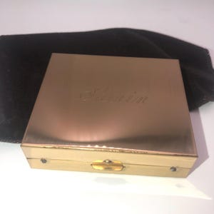 May include: A gold-toned compact with the word "Séduir" engraved on the lid. The compact is rectangular with rounded corners and a gold clasp. A black velvet pouch is behind the compact.