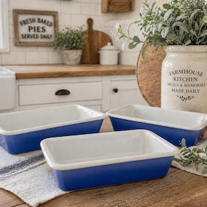 May include: Three rectangular baking dishes with a white interior and a vibrant blue exterior. The dishes are arranged on a white and blue striped cloth. The background includes a farmhouse-style kitchen with a sign that reads "Fresh Baked Pies Served Daily."