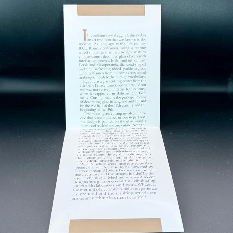 May include: A white rectangular informational card featuring a tan horizontal bar at the top and bottom. The card contains printed text detailing the history and traditional process of glass cutting, starting with the words This brilliant crystal egg.