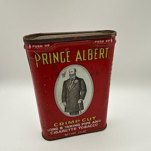 May include: A vintage red Prince Albert tobacco tin. The tin features the text "Prince Albert" in yellow, with an illustration of a man in a suit. The text "Crimp Cut Long Burning Pipe and Cigarette Tobacco" is also visible. Net weight is 1 3/8 oz.