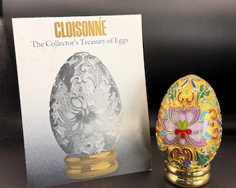 May include: A decorative cloisonné egg with a gold base, featuring a floral design in purple, red, and green against a gold background. The egg is next to a card that reads "CLOISONNÉ The Collector's Treasury of Eggs."