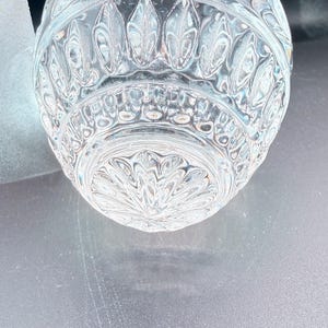 May include: Close-up of a clear, cut-glass object with a detailed, symmetrical pattern. The design features repeating leaf-like shapes and intricate floral motifs. The glass has a reflective quality, catching the light and creating a sparkling effect.