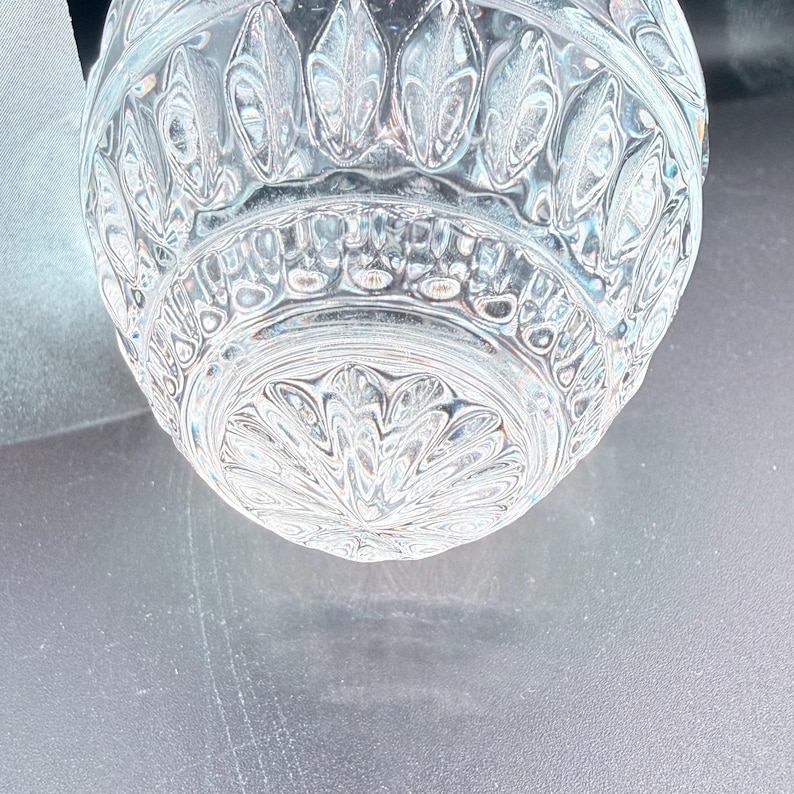 May include: Close-up of a clear, cut-glass object with a detailed, symmetrical pattern. The design features repeating leaf-like shapes and intricate floral motifs. The glass has a reflective quality, catching the light and creating a sparkling effect.