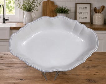 May include: A white, oval-shaped serving platter with a scalloped edge and a subtle blue trim. The platter is displayed on a metal stand, showcasing its elegant design. The kitchen setting includes a wooden cutting board and a sign that reads "FARM SWEET FARM."