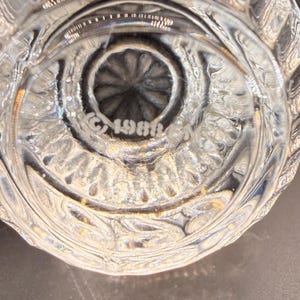 May include: Close-up of a clear, ornate glass object. The base features a circular design with a central, textured pattern. The text "&copy;1986" is etched into the glass. The glass has a detailed, textured pattern.