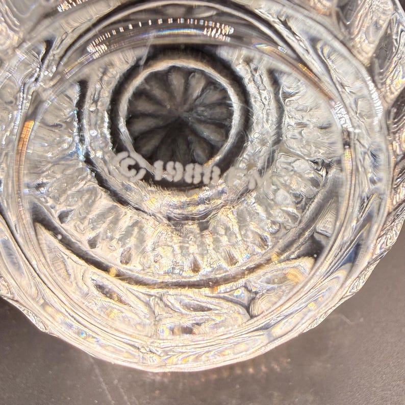 May include: Close-up of a clear, ornate glass object. The base features a circular design with a central, textured pattern. The text "&copy;1986" is etched into the glass. The glass has a detailed, textured pattern.
