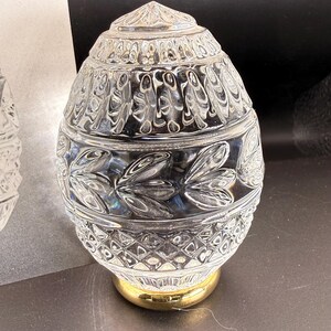 May include: A clear, egg-shaped glass lamp shade featuring a textured pattern of vertical leaves and a diamond-cut base. The glass shade is mounted on a smooth, polished gold-toned metal circular base, creating a reflective and decorative piece.