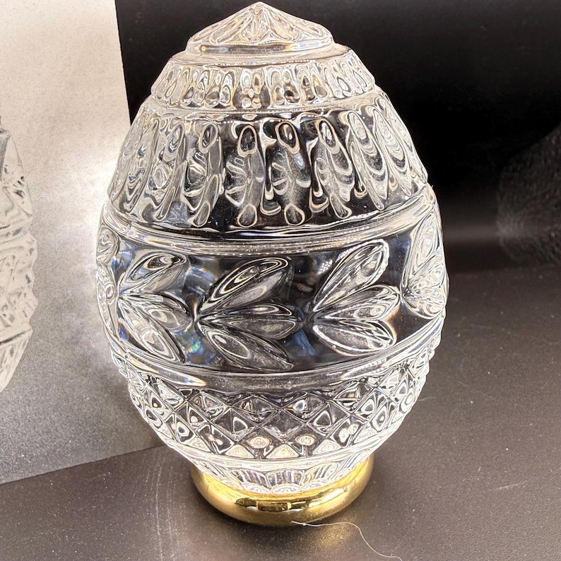 May include: A clear, egg-shaped glass lamp shade featuring a textured pattern of vertical leaves and a diamond-cut base. The glass shade is mounted on a smooth, polished gold-toned metal circular base, creating a reflective and decorative piece.