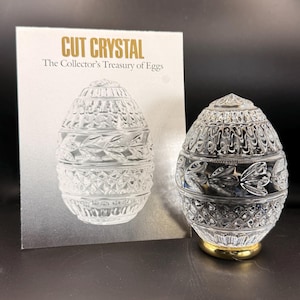 May include: A clear cut crystal egg with intricate leaf and diamond patterns, resting on a gold-colored base. The egg is next to a card that reads "CUT CRYSTAL The Collector's Treasury of Eggs."