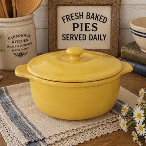 May include: A bright yellow ceramic casserole dish with a lid, handles, and a knob. The dish sits on a beige and blue striped cloth. A sign in a wooden frame reads "FRESH BAKED PIES SERVED DAILY".