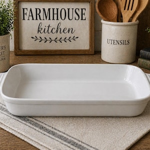 May include: A rectangular white ceramic baking dish with handles, set on a striped linen cloth. The background includes a sign that reads "FARMHOUSE kitchen" and a container labeled "UTENSILS".