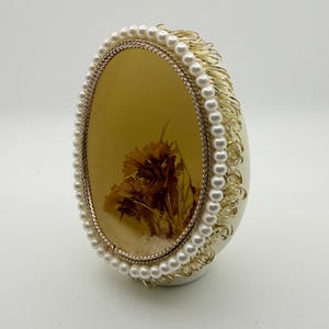 May include: An oval-shaped decorative object with a cream-colored base. The center features dried flowers encased in a clear resin. The resin is framed by a gold-colored metal and a border of faux pearls. The sides have gold-colored filigree.