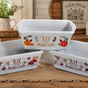 May include: Three white ceramic loaf pans with a rectangular shape and handles. Each pan is decorated with fall-themed graphics, including pumpkins, leaves, and the text "Fall is my favorite color."