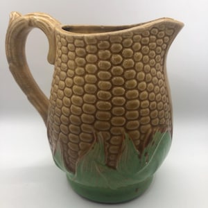 Könnte beinhalten: Eine Keramikkrug mit einem Maiskolben-Design. Der Krug ist beige mit einer strukturierten Oberfläche, einem braunen Griff und grünen Blättern am Boden. Der Krug ist zum Halten von Flüssigkeiten bestimmt.
