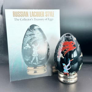 以下が含まれることがあります： 2つの装飾的なロシア漆器スタイルの卵。1つの卵は、赤いドラゴンのデザインと青い人物が描かれた黒色です。もう1つの卵は、人物とドラゴンのデザインが施された灰色です。カードのテキストは「Russian Lacquer Style The Collector's Treasury of Eggs」と書かれています。