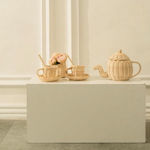Peut inclure: Un service à thé en osier clair, comprenant une théière, deux tasses avec soucoupes et un petit panier de fleurs roses. La théière a un bec courbé et un couvercle. Les tasses ont des anses et reposent sur des soucoupes assorties. L'ensemble est en osier tressé.