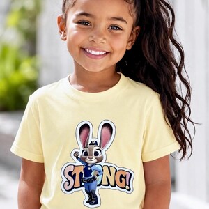 Op de afbeelding: Een lichtgeel t-shirt met een cartoonafbeelding van Judy Hopps uit Zootopia, met het woord "STRONG!" in kleurrijke letters. Het shirt wordt gedragen door een jong persoon met lang donker haar en een spijkerbroek.