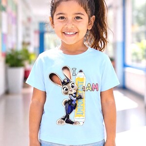 K&ouml;nnte beinhalten: Hellblaues T-Shirt mit einem Cartoon-Hasen in Polizeiuniform, der einen Bleistift mit der Aufschrift "I AM IMPORTANT" h&auml;lt. Das T-Shirt wird von einem jungen M&auml;dchen getragen. Das M&auml;dchen tr&auml;gt Jeansshorts.