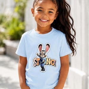 Op de afbeelding: Een lichtblauw T-shirt met een cartoonafbeelding van Judy Hopps uit Zootopia, met het woord "STRONG!" in een kleurrijk lettertype. Het T-shirt wordt gedragen door een jong meisje met lang donker haar.