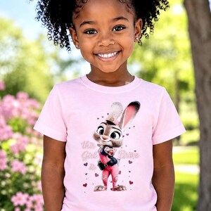 May include: A young girl wearing a light pink t-shirt featuring a cartoon bunny character in a police uniform. The shirt has the text "This Girl is Brave" with heart accents. The girl is smiling, with curly hair and wearing a pink bow.