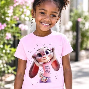 May include: A pink t-shirt featuring a cartoon bunny wearing glasses and a fanny pack, with the text "DON'T WORRY BE HOPPY!". The shirt is worn by a young girl with curly hair.