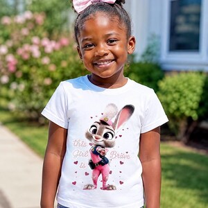 May include: A young girl wearing a white t-shirt featuring a cartoon bunny character in pink attire. The shirt has the text "This Girl is Brave" and heart graphics. She is also wearing a pink bow in her hair.