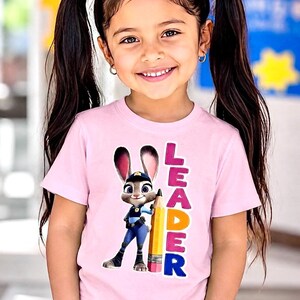 May include: A light pink t-shirt with a cartoon rabbit police officer holding a pencil, and the word "LEADER" in colourful letters. The shirt is worn by a young girl with dark hair in pigtails.