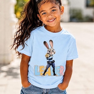 May include: A young girl wearing a light blue t-shirt with a cartoon image of Judy Hopps from Zootropolis, holding a star badge. The word "LEADER" is printed below the image in colourful letters. She is also wearing denim shorts.