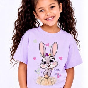 May include: A light purple short-sleeved t-shirt featuring a graphic of a grey cartoon rabbit wearing a yellow skirt and a flower crown. The text "Pretty Little Bunny" is printed in purple script alongside small pink and light green hearts.