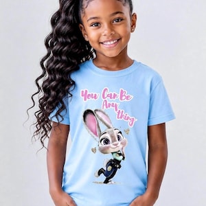 May include: A light blue t-shirt featuring a cartoon image of Judy Hopps from Zootopia. The shirt has the text "You Can Be Anything" in pink script. The girl is wearing blue jeans.