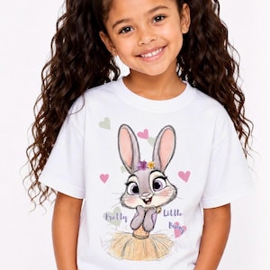 May include: A white short-sleeved t-shirt featuring a graphic illustration of a grey rabbit with large purple eyes, wearing a yellow skirt and floral crown, surrounded by small hearts and the text Pretty Little Bunny in purple script.