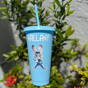 May include: A light blue plastic tumbler with a matching lid and straw. The cup features the white text KAELANY printed near the top and a graphic of a grey and white rabbit character wearing a blue police uniform centred on the front.