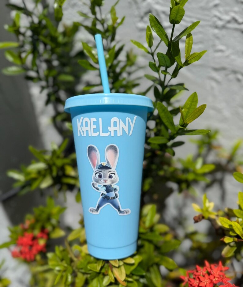 May include: A light blue plastic tumbler with a matching lid and straw. The cup features the white text KAELANY printed near the top and a graphic of a grey and white rabbit character wearing a blue police uniform centred on the front.