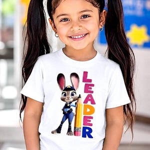 May include: A young girl wearing a white t-shirt featuring a cartoon bunny holding a pencil, with the word "LEADER" in pink, orange, and blue. The girl is smiling and wearing blue jeans.