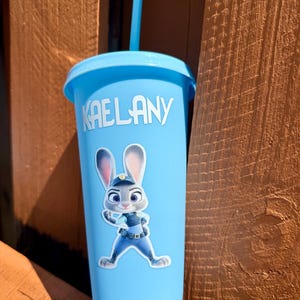 May include: A light blue plastic tumbler with a matching lid and straw. The cup features the white text KAELANY and a graphic of a cartoon rabbit character wearing a blue police uniform with a badge and utility belt on the front surface.