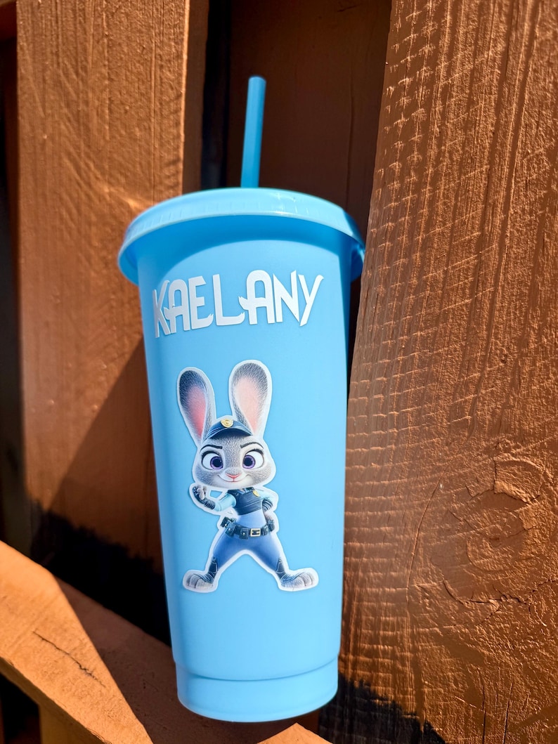 May include: A light blue plastic tumbler with a matching lid and straw. The cup features the white text KAELANY and a graphic of a cartoon rabbit character wearing a blue police uniform with a badge and utility belt on the front surface.