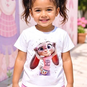 May include: A young girl smiles, wearing a white t-shirt featuring a cartoon bunny character with glasses and the text "Don't Worry Be Hoppy!". She also wears pink shorts and has pink bows in her hair.