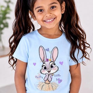 May include: A light blue short-sleeved t-shirt featuring a graphic illustration of a grey rabbit with large eyes, wearing a floral crown and a tan skirt. The text "Pretty Little Bunny" is printed in script next to the rabbit, surrounded by small purple and blue hearts.