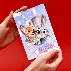 May include: A light blue greeting card featuring cartoon characters Nick Wilde and Judy Hopps from Zootopia. The card has the text "TO: FROM:" at the top and "FRIENDS FUR LIFE" at the bottom. The card is held against a red background.