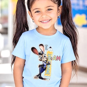 K&ouml;nnte beinhalten: Ein junges M&auml;dchen mit dunklem Haar in Z&ouml;pfen l&auml;chelt und tr&auml;gt ein hellblaues T-Shirt. Das Shirt zeigt ein Cartoon-Bild eines Hasen in Polizeiuniform, der einen Bleistift mit den Worten "I AM IMPORTANT" h&auml;lt.